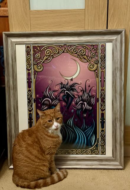 Fred, an orange tabby cat, sitting on the carpet in front of a framed print of Oscar’s Garden
