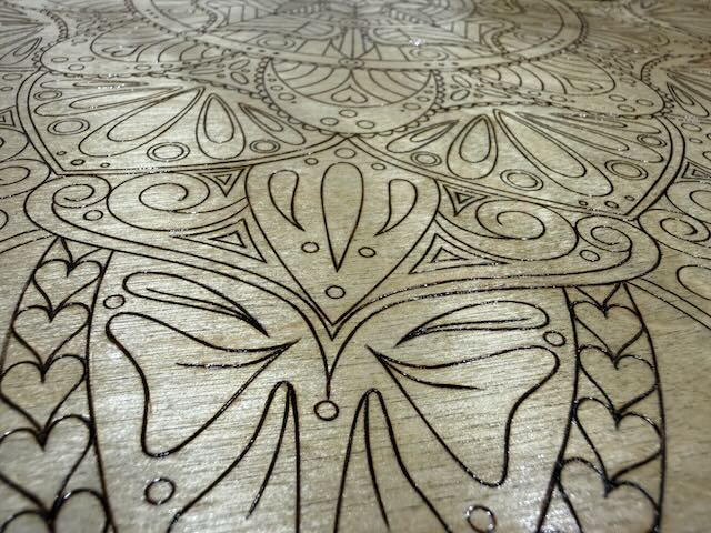 Close-up of the Hearty Mandala being hand burnt into pale wood, showing outlined petals, teardrops and a row of small hearts around the outer band